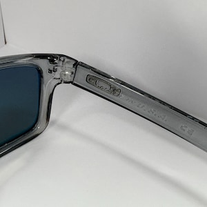 May include: Gray sunglasses with blue lenses. The frame is a translucent gray color. The text "MADE IN U.S.A." is printed on the arm of the glasses.