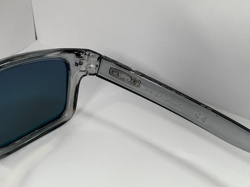 May include: Gray sunglasses with blue lenses. The frame is a translucent gray color. The text "MADE IN U.S.A." is printed on the arm of the glasses.