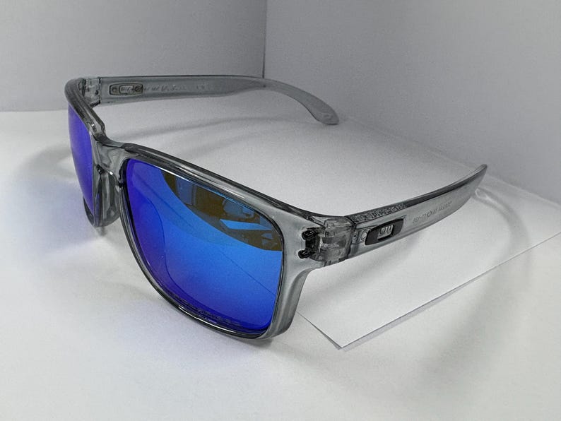 May include: Gray sunglasses with blue lenses. The frames are a translucent gray, and the lenses reflect a bright blue hue. The sunglasses have a rectangular shape and a modern design, suitable for outdoor activities.