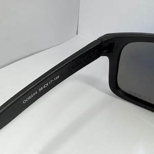 May include: Black sunglasses with a rectangular frame. The arm of the sunglasses has the text "OO9244 56 17-138". The lenses have a gradient tint. The sunglasses are on a white surface.