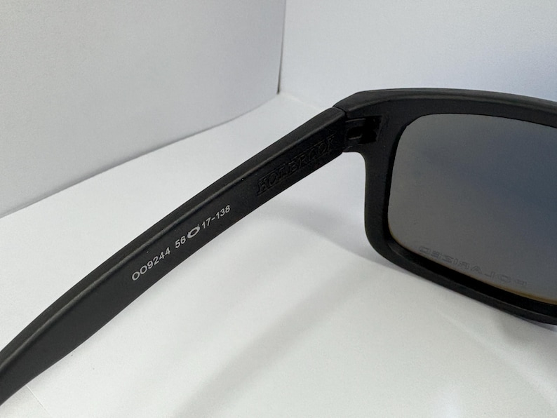 May include: Black sunglasses with a rectangular frame. The arm of the sunglasses has the text "OO9244 56 17-138". The lenses have a gradient tint. The sunglasses are on a white surface.