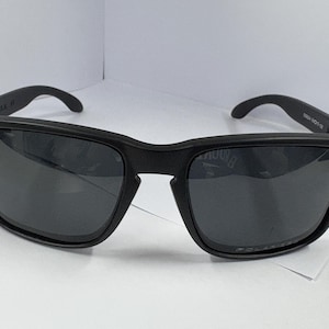 May include: Black sunglasses with a square frame. The sunglasses have dark lenses and a matte finish. The arms of the sunglasses curve around the ears. The sunglasses are on a white surface.