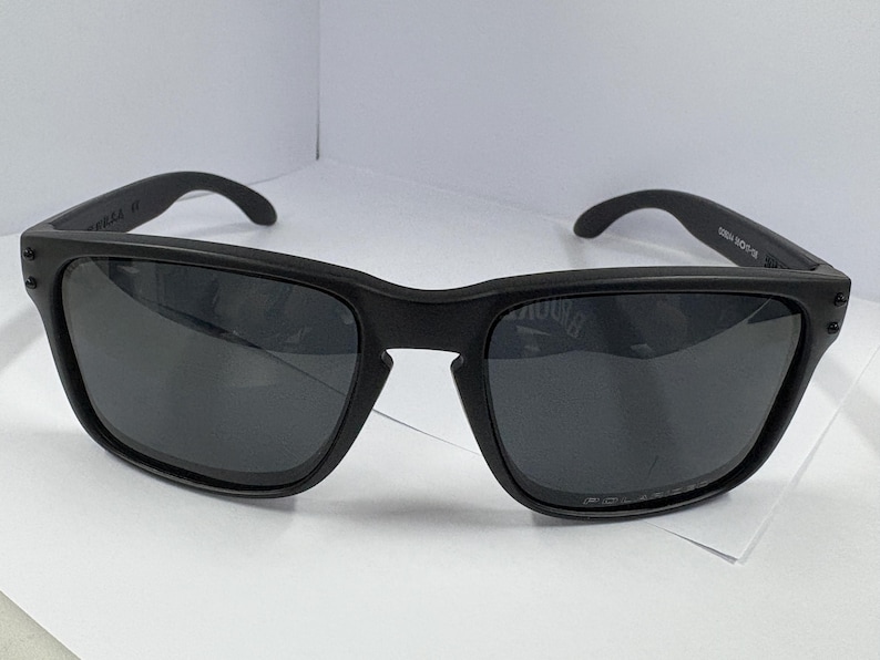 May include: Black sunglasses with a square frame. The sunglasses have dark lenses and a matte finish. The arms of the sunglasses curve around the ears. The sunglasses are on a white surface.