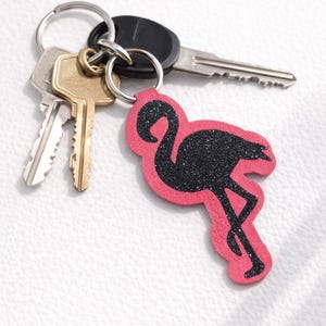 May include: A key ring with a pink flamingo-shaped charm with black glitter. The key ring holds several keys, including a car key and house keys. The flamingo charm is a decorative accessory.