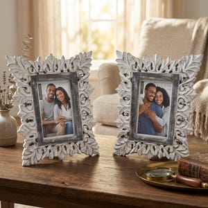 Puede incluir: Dos marcos de fotos decorativos con acabado blanco envejecido y tallas ornamentales en forma de hojas. Cada marco contiene una fotografía de una pareja. Los marcos están sobre una mesa de madera, para decoración del hogar.