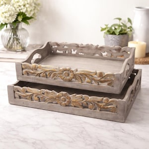 May include: Two decorative wooden trays, one stacked on top of the other. The trays are rectangular with carved floral and leaf designs in a contrasting gold color. The trays are painted in a light gray color.
