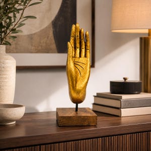 Può includere: Una scultura color oro di una mano aperta con linee del palmo visibili, montata verticalmente su una sottile asta di metallo nero fissata a una base quadrata in legno marrone scuro. La mano è posizionata con le dita rivolte verso l'alto.