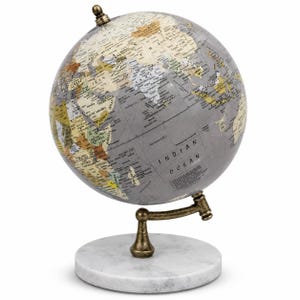 May include: A decorative globe featuring a grey and cream color palette with printed geographical labels. The sphere is mounted on a metallic bronze-toned arm attached to a circular white marble base, displaying the Indian Ocean and surrounding landmasses.