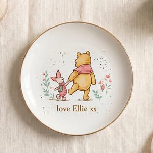 Puede incluir: Plato de cerámica blanco con borde dorado. Presenta una ilustración de Winnie the Pooh y Piglet tomados de la mano, con detalles florales y el texto "love Ellie xx".