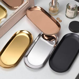 May include: A selection of oval trays in gold, silver, copper, and black. The trays are displayed on a white surface, with a mirror and perfume bottles in the background. These trays are likely for decorative or organizational use.