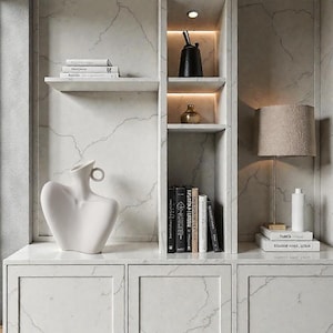 May include: A white cabinet with integrated shelving and a marble-effect background. The shelves hold books, a white vase, a black vase, and a gold ornament. A lamp with a beige lampshade is positioned on the cabinet.