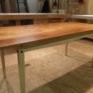 Puede incluir: Una mesa de madera con una tapa de madera clara y patas pintadas de blanco. La mesa tiene un diseño sencillo con patas cuadradas y una tapa rectangular.