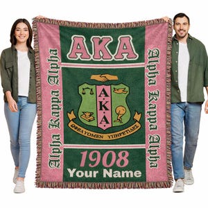 Op de afbeelding: Een geweven sprei in roze, groen en wit, met de letters "AKA" en een wapen. De deken heeft de woorden "Alpha Kappa Alpha" aan de zijkanten en "1908" met ruimte voor een naam onderaan.