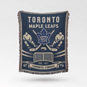 May include: Blue woven throw blanket featuring the Toronto Maple Leafs logo and text. The design includes hockey players, crossed hockey sticks, a net, and the text "EST. 1917 Toronto, Canada". The blanket has a fringed edge.