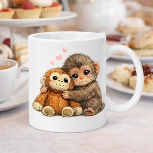 Puede incluir: Taza de cerámica blanca con una ilustración de dibujos animados de dos monos abrazándose, con corazones encima. Un mono es marrón y el otro es marrón claro. La taza tiene un asa curva. Pasteles en el fondo.