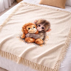 Op de afbeelding: Een crèmekleurige sprei met een aquarel illustratie van twee knuffelende apen. De sprei heeft een gefranjerde rand en ligt op een bed met witte lakens en een beige kussen. De apen zijn bruin en oranje.