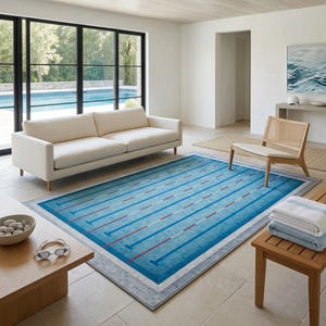 Puede incluir: Una alfombra rectangular con un diseño de carriles de piscina en azul con líneas blancas y rojas, bordeada por un marco gris. La alfombra está colocada sobre un suelo de baldosas claras en una habitación con un sofá blanco y una silla de madera.