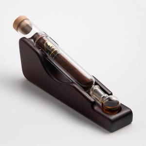 May include: A clear glass tube containing a dark brown cigar with a gold and black label, sealed with a light brown cork stopper. The tube rests at an angle in a dark brown wooden stand with a circular indentation for the glass bulb base.