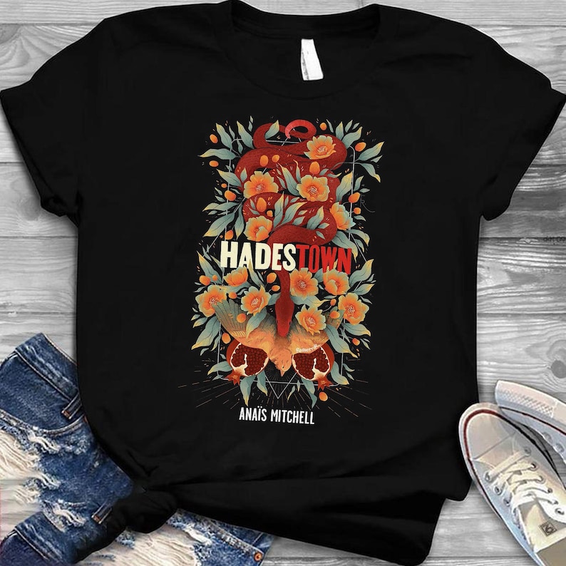 K&ouml;nnte beinhalten: Schwarzes T-Shirt mit einem farbenfrohen Grafikdesign. Das Design zeigt den Text "HADESTOWN" in Wei&szlig; und Rot, eine Schlange, Blumen und den Text "ANAIS MITCHELL". Das Shirt besteht aus einem dunklen Stoff.