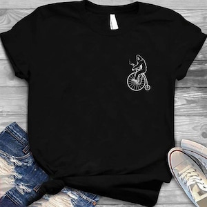 Peut inclure: T-shirt noir avec un dessin blanc d'une personne sur un grand-bi fumant. Le motif est sur la poitrine gauche. Le t-shirt est présenté sur une surface en bois avec un jean et des baskets.