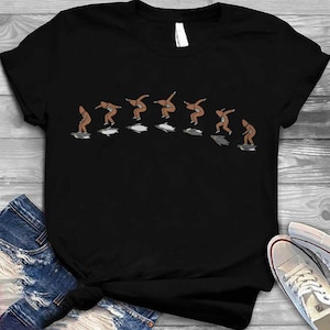 Peut inclure: T-shirt noir avec un motif représentant une silhouette surfant sur des vagues stylisées. La silhouette est représentée dans une séquence de poses, illustrant l'action du surf. Le design utilise des couleurs marron et blanc.