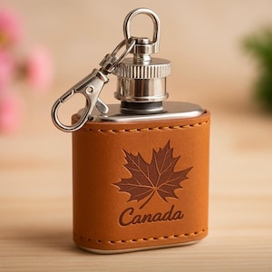 Op de afbeelding: Een bruin lederen heupfles met een zilveren dop en een karabijnhaak. De fles heeft een esdoornbladontwerp en het woord "Canada" gegraveerd op de voorkant. De fles is ontworpen om vloeistoffen te bevatten.