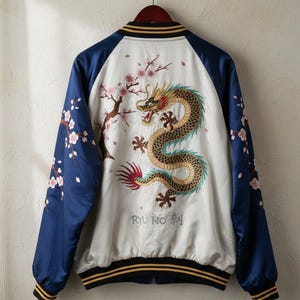 May include: A satin bomber jacket with a white body and navy blue sleeves. The back features an embroidered golden dragon, cherry blossoms, and Japanese text. The jacket has black and gold striped trim at the collar, cuffs, and hem.