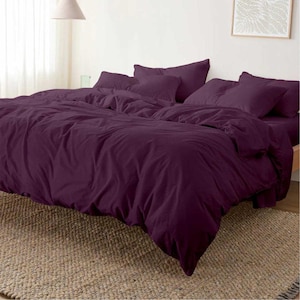 Puede incluir: Un juego de cama de color púrpura oscuro, que incluye una funda nórdica y varias almohadas, colocado sobre una cama. La ropa de cama tiene un aspecto suave y ligeramente arrugado. La cama está sobre una alfombra tejida.