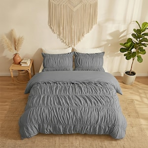 May include: A gray bedspread and two matching pillowcases with a ruched design are displayed on a bed. A woven wall hanging and a small side table with a vase and books are visible. A potted plant is on the right.