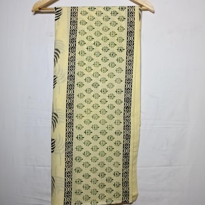 Puede incluir: Tela de color crema con un estampado floral verde repetido y un borde negro. La tela está colgada de una percha de madera. El diseño incluye un patrón repetido de líneas curvas negras en el lado izquierdo.