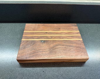 May include: A square wooden cutting board with a rich brown finish. The board features three parallel, lighter-colored wood inlays. The wood grain is visible, and the board appears to be handcrafted. The cutting board is approximately 12 inches square.