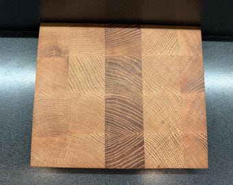 May include: A square, end-grain butcher block cutting board. The board is made of light and dark brown wood blocks arranged in a checkerboard pattern. The wood grain is visible, adding texture to the surface.