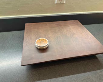 May include: A large, square, end-grain cutting board made of dark wood. The board has a rich, reddish-brown color and a circular grain pattern. A small, round tin of cutting board oil sits on the surface.