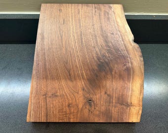 May include: A rectangular, live edge wooden board with a natural, dark brown finish. The wood grain is visible, and the board has a smooth surface. The board is likely used for serving or display.