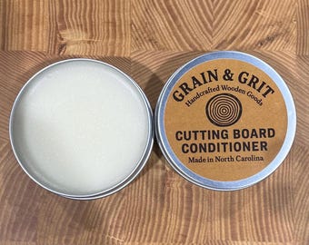 May include: Two small, round metal tins of cutting board conditioner. One tin is open, revealing a solid, white substance. The other tin is closed, with a brown label that reads "Grain & Grit Handcrafted Wooden Goods CUTTING BOARD CONDITIONER Made in North Carolina."