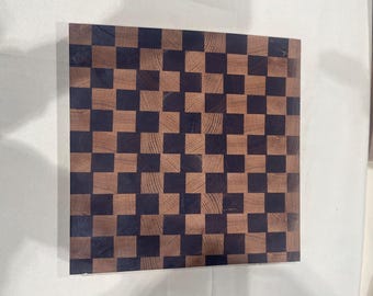 May include: A square wooden cutting board with a checkerboard pattern. Alternating squares are dark brown and light brown, creating a visually appealing design. The board is made of wood and has a smooth surface.