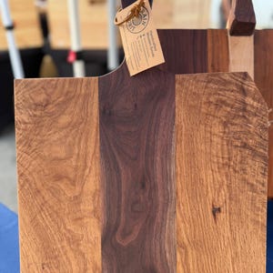 Könnte beinhalten: Ein rechteckiges Servierbrett aus Holz mit einem dunkelbraunen, vertikalen Mittelstreifen, der von zwei hellbraunen Holzpaneelen mit sichtbarer Maserung flankiert wird. Am Griff befindet sich ein beiges Etikett mit der Aufschrift Grain Grit.
