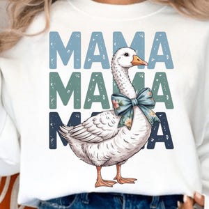 Op de afbeelding: Witte sweatshirt met een ganzenillustratie en een blauwe bloemenstrik. Het woord "MAMA" is drie keer in blauw en blauwgroen gestapeld. Een casual en comfortabele top.