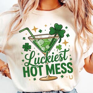 May include: A cream-colored t-shirt featuring a green cocktail with ice cubes, a straw, and a shamrock. The text "Luckiest HOT MESS" is written in green. Gold stars and shamrocks decorate the design, with a horseshoe at the bottom.