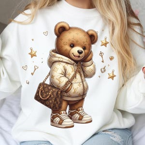 May include: White sweatshirt featuring a cartoon bear wearing a hoodie, carrying a purse, and wearing sneakers. The bear is brown with a designer pattern. Gold stars, keys, and hearts are scattered around the bear.