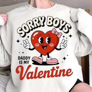 Puede incluir: Sudadera blanca con el texto "SORRY BOYS" sobre un personaje de corazón de dibujos animados. El corazón tiene un lazo rosa, brazos, piernas y hace signos de paz. El texto "DADDY IS MY Valentine" está debajo.
