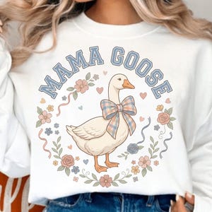 Könnte beinhalten: Weißes Sweatshirt mit einer Grafik einer Gans mit karierter Schleife, umgeben von einem floralen Kranz. Der Text "MAMA GOOSE" ist in blauen Buchstaben über der Gans angeordnet. Das Design beinhaltet Herzen und pastellfarbene Blumen.