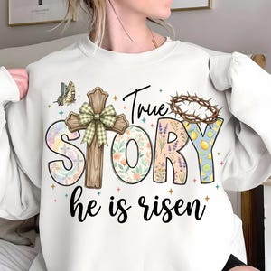 May include: White sweatshirt with the words "True Story he is risen" in a colorful design. The design includes a cross, floral patterns, a crown of thorns, and a butterfly. The sweatshirt is a casual, comfortable style.