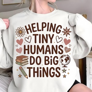 Op de afbeelding: Witte sweatshirt met de tekst "HELPING TINY HUMANS DO BIG THINGS" in bruin. Het ontwerp bevat een bloem, harten, boeken, een wereldbol, een ster en een potlood. Een comfortabele top voor leraren of kinderopvang.