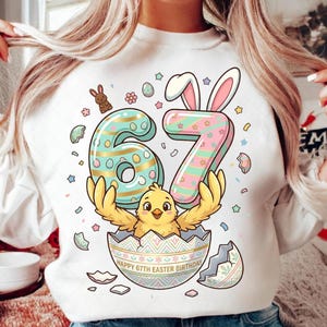 Könnte beinhalten: Weißes Sweatshirt mit einem farbenfrohen Oster-Motiv. Das Design zeigt die Zahl 67 mit Hasenohren, ein Küken, das aus einem Ei schlüpft, und den Text "HAPPY 67TH EASTER BIRTHDAY". Die Grafik enthält Pastellfarben und Osterei-Details.