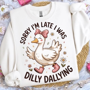 May include: White sweatshirt with a cartoon duck wearing a pink bow and boots, with the text "SORRY I'M LATE I WAS DILLY DALLYING". The duck is walking with a smile, surrounded by flowers and confetti.