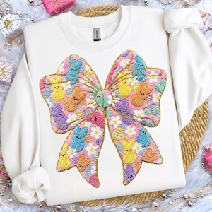 Könnte beinhalten: Weißes Sweatshirt mit einem großen, gestickten Schleifendesign. Die Schleife ist mit bunten Elementen zum Thema Ostern gefüllt, darunter Hasen, Eier und Blumen. Die Schleife hat eine goldene Umrandung. Ein festliches und fröhliches Kleidungsstück. Ein bequemes Kleidungsstück.