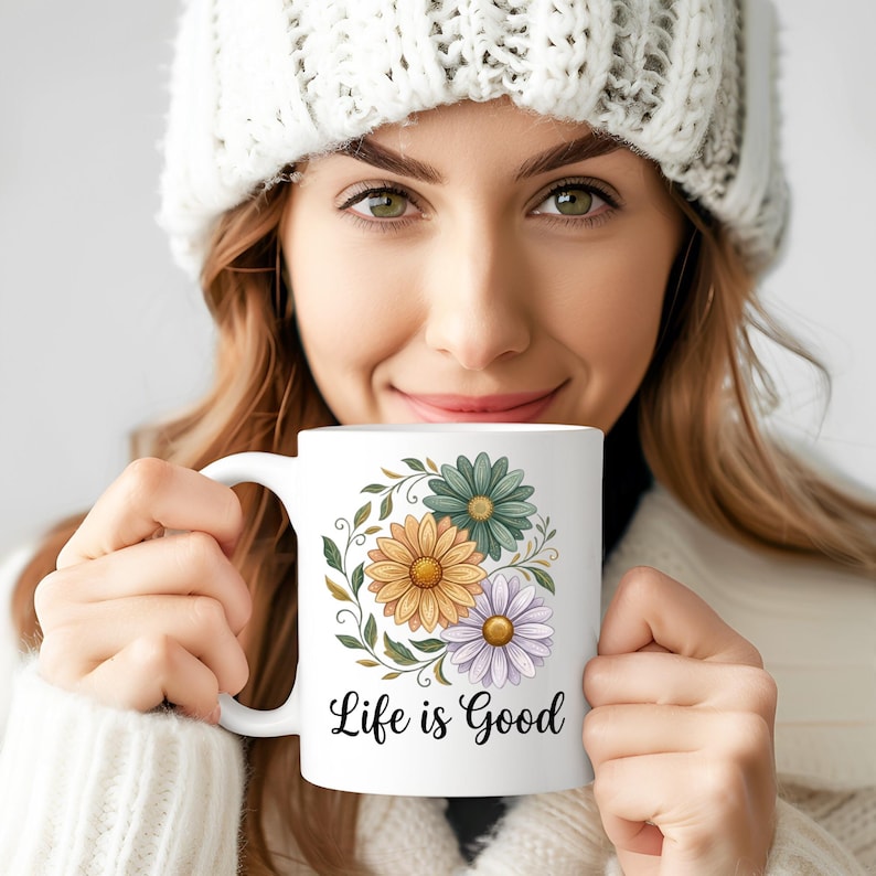 Puede incluir: Taza de cer&aacute;mica blanca con un dise&ntilde;o floral de flores amarillas, verdes y moradas, y el texto "Life is Good". La taza tiene un asa curva. La persona que sostiene la taza lleva un gorro y un su&eacute;ter blancos.