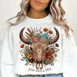 May include: White sweatshirt featuring a detailed illustration of a bull skull adorned with flowers, feathers, and turquoise accents. The skull has intricate patterns, and the text "Stay Wild & Free" is printed at the bottom. A bohemian-style design.