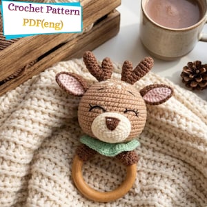 Könnte beinhalten: Ein gehäkeltes Rentier-Rassel mit einem Holzring-Sockel. Das Rentier hat braunes und cremefarbenes Garn mit einem grünen Kragen. Der Text "Crochet Pattern PDF(eng)" befindet sich in der oberen linken Ecke.